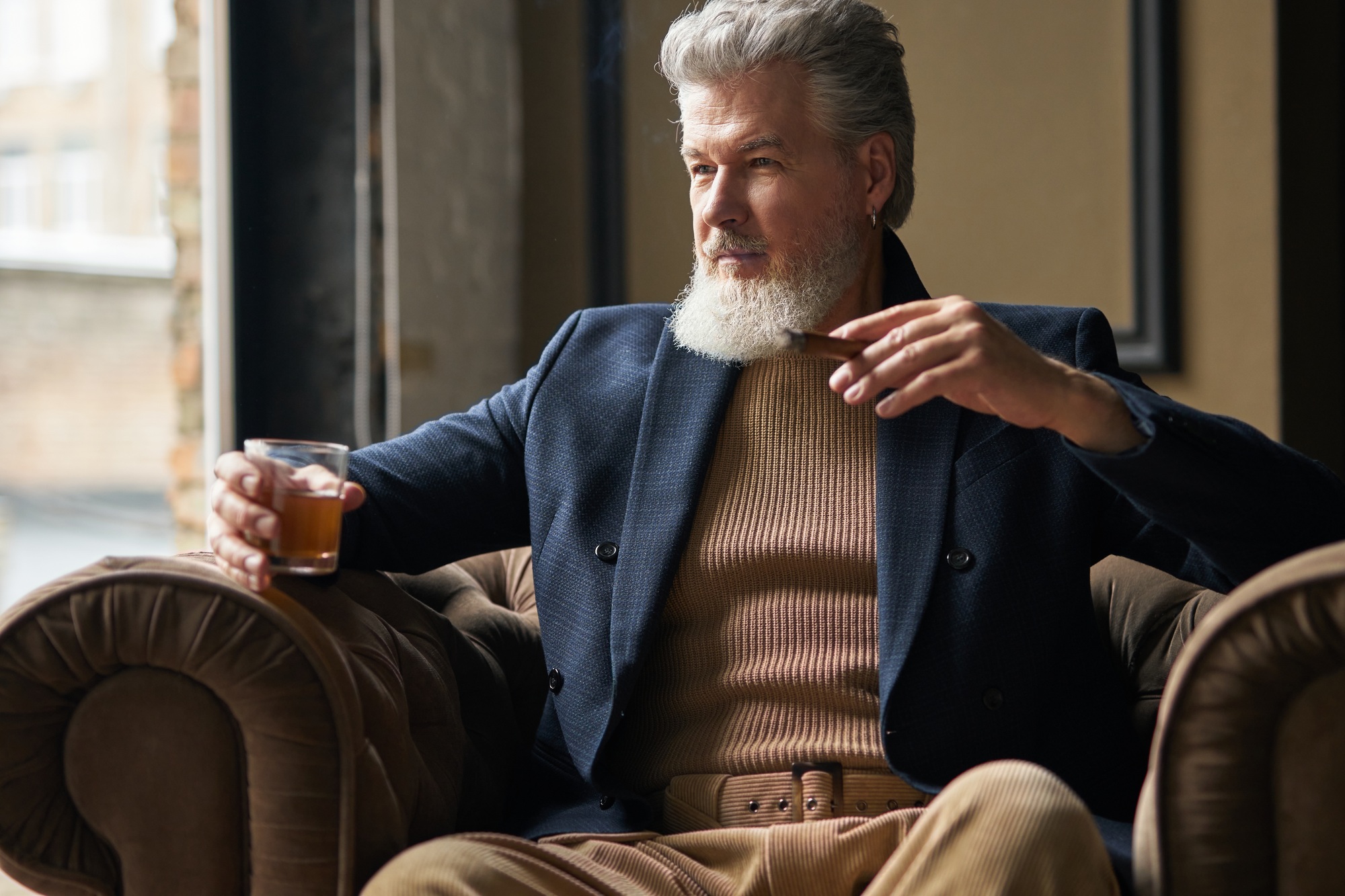 Portrait of elegant mature businessman with beard looking aside while holding glass of whisky and